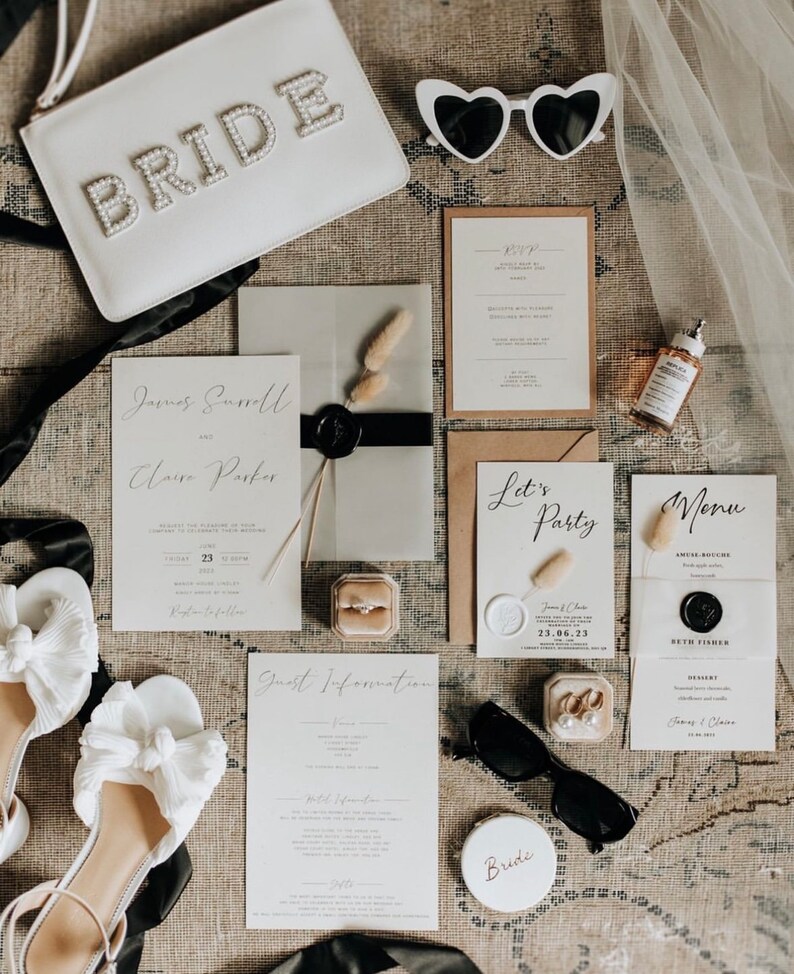 May include: A flat lay of wedding accessories including a white clutch with "BRIDE" in pearl lettering, heart-shaped sunglasses, invitations, a ring box, and white bow sandals. The invitations feature elegant calligraphy and a wax seal.