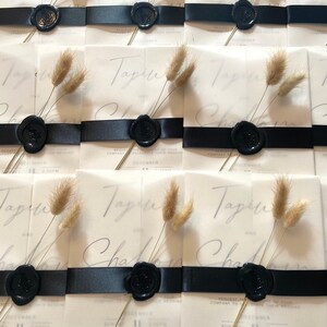 May include: Wedding invitations with a translucent overlay, featuring the names "Tapin" and "Charlie" in elegant script. Each invitation is adorned with a black ribbon, a black wax seal, and a sprig of dried bunny tail grass. The date "December 11" is visible.