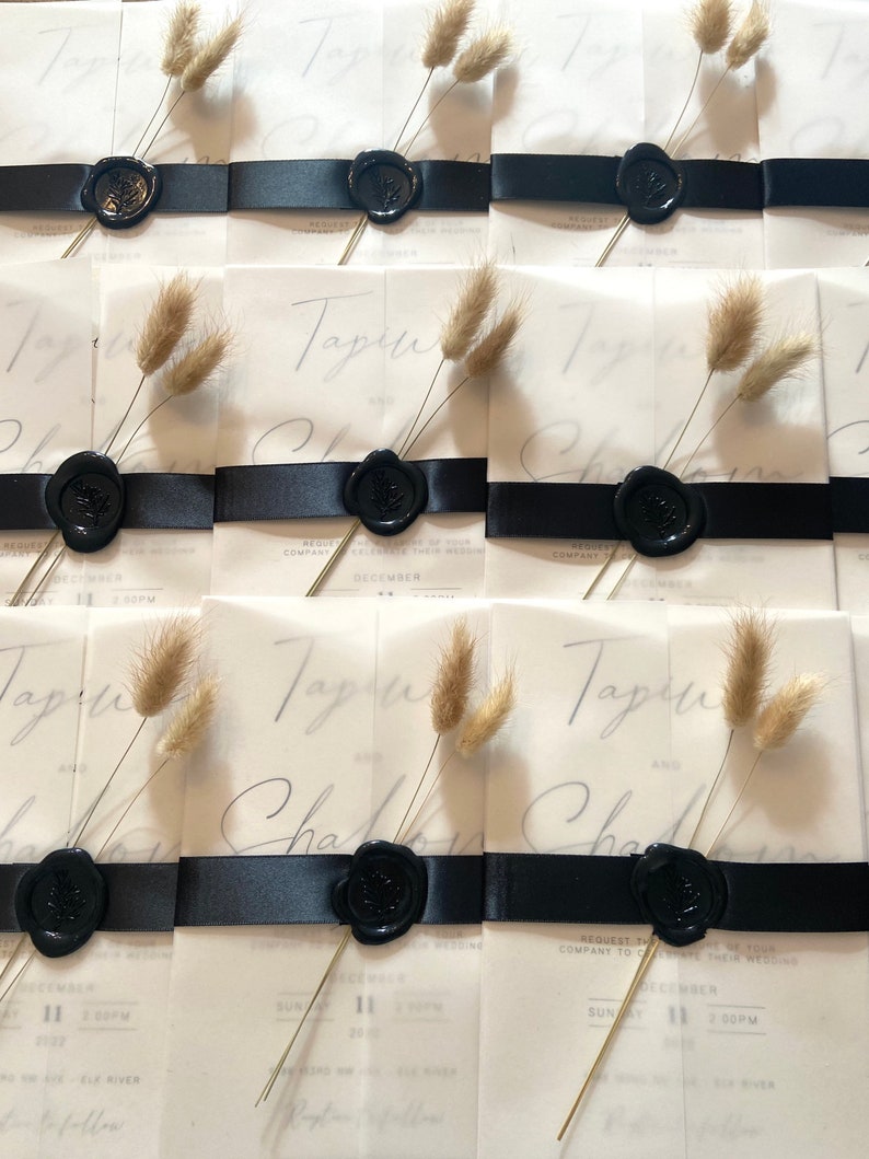 May include: Wedding invitations with a translucent overlay, featuring the names "Tapin" and "Charlie" in elegant script. Each invitation is adorned with a black ribbon, a black wax seal, and a sprig of dried bunny tail grass. The date "December 11" is visible.