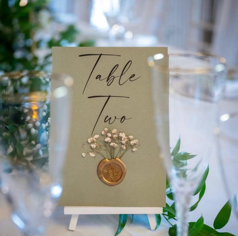 May include: A green card with the text "Table Two" in black calligraphy. A small bouquet of white flowers is attached to the card with a gold wax seal.