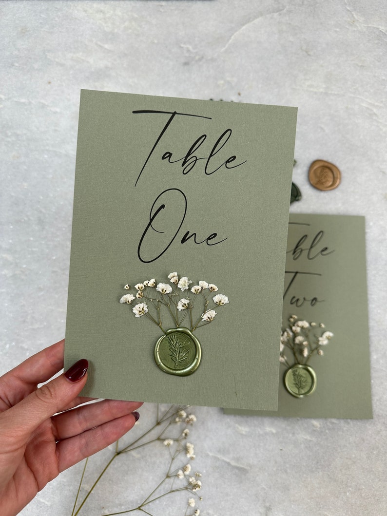 May include: Green table number card with the text "Table One" in black calligraphy. The card is decorated with white baby's breath flowers and a green wax seal.