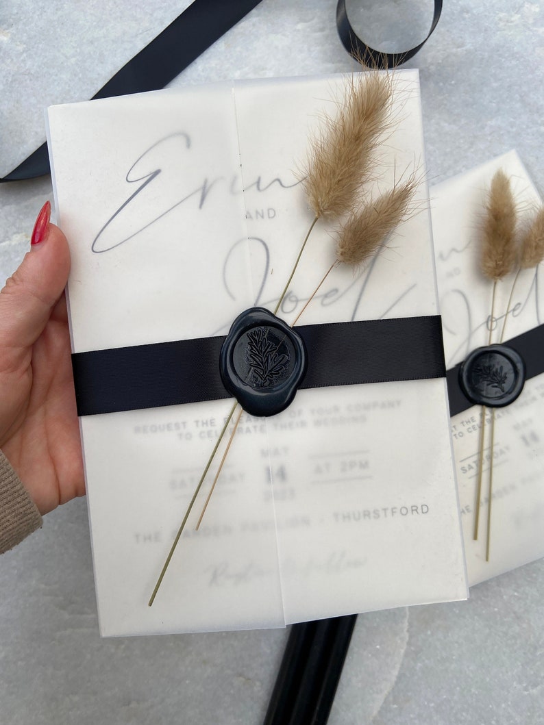 May include: Wedding invitation suite featuring a translucent vellum overlay with the names "Erin and Joel" in elegant script. The invitation is secured with a black ribbon and a dark blue wax seal, accented with dried bunny tail grass.