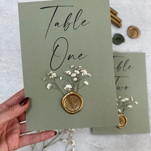 May include: Sage green card with the text "Table One" in black calligraphy. A gold wax seal with a leaf design is on top of a cluster of white baby's breath flowers.