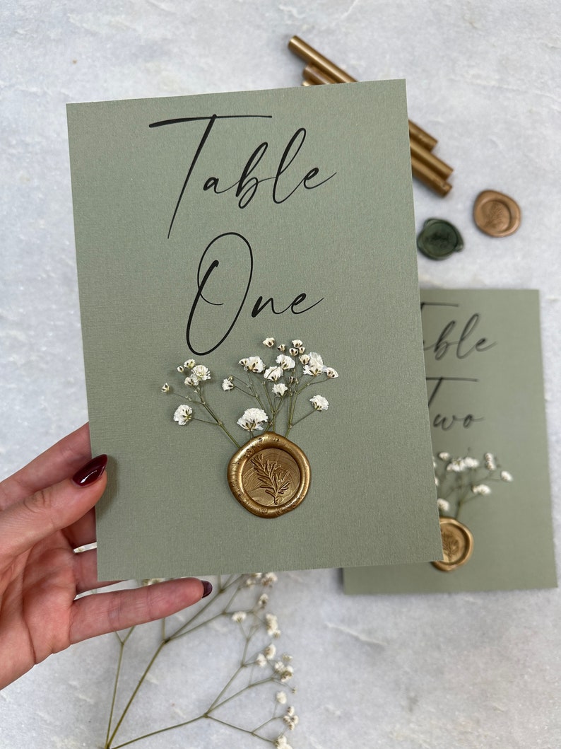 May include: Sage green card with the text "Table One" in black calligraphy. A gold wax seal with a leaf design is on top of a cluster of white baby's breath flowers.