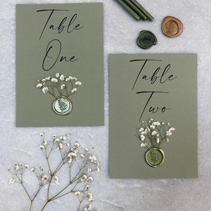 May include: Two green table number cards with white flowers and wax seals. The cards say "Table One" and "Table Two".