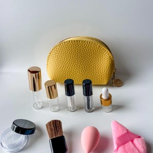 May include: A collection of beauty products and accessories, including small glass bottles with gold and black caps, a yellow textured cosmetic bag, a makeup brush, a pink sponge, and a powder puff.