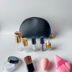 May include: A black leather cosmetic bag with a variety of beauty products. Includes small glass bottles with gold and black caps, a makeup brush, a pink sponge, and a powder puff.