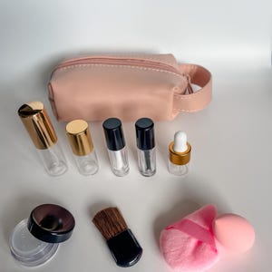 May include: A collection of beauty products, including clear glass bottles with gold and black caps, a small pink cosmetic bag, a makeup brush, a powder puff, and a makeup sponge. The items are arranged on a white surface.