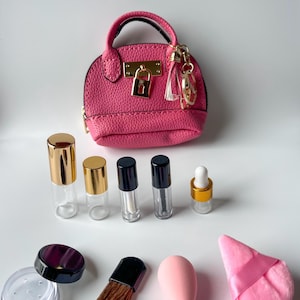 Pode incluir: Uma coleção de produtos e acessórios de beleza, incluindo uma bolsa rosa, pequenos frascos de vidro com tampas douradas e pretas, um pincel de maquiagem, uma esponja rosa e uma borla. Os itens estão dispostos em uma superfície branca.