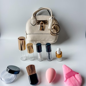 Puede incluir: Un conjunto de productos y accesorios de belleza, que incluye un pequeño bolso blanco con detalles dorados, botellas de vidrio transparente con tapas doradas y negras, un pincel de maquillaje, una esponja rosa y una borla rosa.