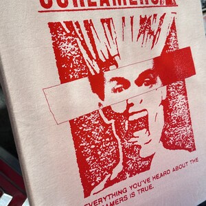 Puede incluir: Una camiseta blanca con un estampado gr&aacute;fico rojo de una persona con el pelo de punta y una banda roja que le cubre los ojos. El texto "Screamers" est&aacute; impreso en rojo en la parte superior del gr&aacute;fico y el texto "Everything you've heard about the Screamers is true." est&aacute; impreso en rojo en la parte inferior del gr&aacute;fico.