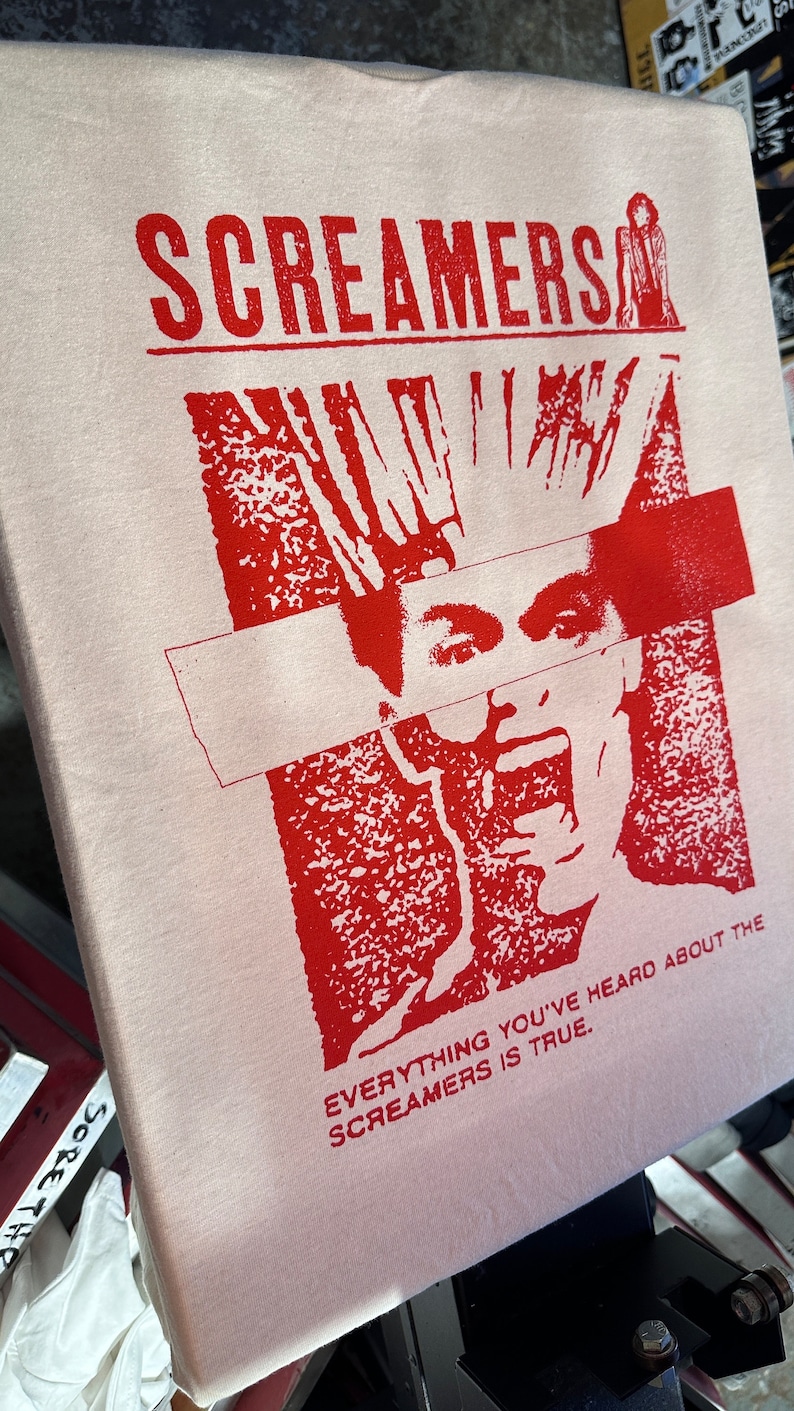Puede incluir: Una camiseta blanca con un estampado gr&aacute;fico rojo de una persona con el pelo de punta y una banda roja que le cubre los ojos. El texto "Screamers" est&aacute; impreso en rojo en la parte superior del gr&aacute;fico y el texto "Everything you've heard about the Screamers is true." est&aacute; impreso en rojo en la parte inferior del gr&aacute;fico.