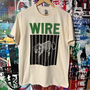 May include: Cream-coloured t-shirt featuring the word "WIRE" in green block letters above a black and white graphic of a cheetah behind vertical black bars. The shirt is hanging on a wooden hanger.