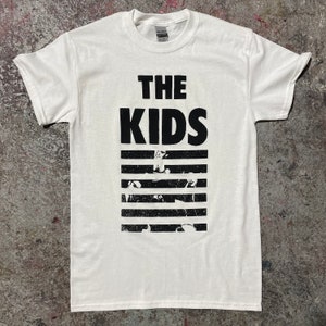 May include: White t-shirt featuring the text "THE KIDS" in bold black letters above a series of horizontal black stripes. The stripes contain faded black and white images. The shirt is made of a soft material.