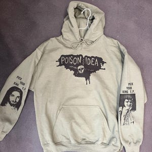 May include: An olive green hooded sweatshirt with the text "POISON IDEA" above a skull graphic. The sleeves feature the text "PICK YOUR KING E.P." along with black and white images. A front pocket is visible.