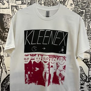 May include: White t-shirt with a black box that says "KLEENEX" and a graphic of Pac-Man and a stick figure. Below the box is a graphic of five cartoon faces with red outlines.