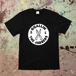 Op de afbeelding: Zwart T-shirt met een wit rond logo met de tekst "GGALLIN & THE JABBERS" en twee gekruiste messen.