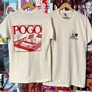 Op de afbeelding: Twee crèmekleurige t-shirts hangen aan houten hangers. Eén heeft een rode afbeelding op de achterkant met het woord "POGO" en informatie over zeefdrukken. De andere heeft een kleine cirkelvormige afbeelding op de voorkant met een persoon en "POGO PRINTING".