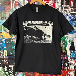 Könnte beinhalten: Schwarzes T-Shirt mit weißem Aufdruck. Der Aufdruck zeigt das Wort "NEANDERTHAL" über dem Text "WEST COAST POWER VIOLENCE" und eine Schwarz-Weiß-Illustration einer Figur. Das T-Shirt hängt an einem Holzbügel.