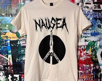 May include: A cream colored t-shirt with a black peace sign graphic and the word "NAUSEA" in black metal font.