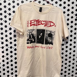 Op de afbeelding: Crèmekleurig T-shirt met de bandnaam "The Ejected" in rood, boven drie zwart-wit afbeeldingen. Onder de afbeeldingen staat de tekst "Have You Got 10p?" in rood gedrukt. Het T-shirt hangt aan een metalen hanger.