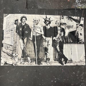 May include: Black and white poster depicting four people in punk rock clothing. The poster includes text and a dog. The people are standing in front of a wall. The poster is on a dark surface.