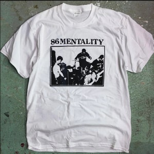 Puede incluir: Camiseta blanca con un estampado gráfico en blanco y negro de una banda tocando música. El texto "86MENTALITY" está impreso sobre el gráfico.