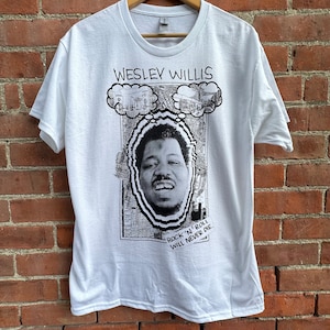 May include: White t-shirt with a black and white graphic of Wesley Willis. The design includes a portrait, thought bubbles, and the text "WESLEY WILLIS" above the image. The text "ROCK 'N' ROLL WILL NEVER DIE" is also present.