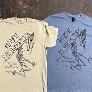 May include: Two t-shirts, one light beige and one light blue, featuring a skeleton graphic holding a flag. The text "HATED PRINCIPLES" is above the graphic, with "THE CURSE OF PRINCE HATED" and "5-SONGS" below.