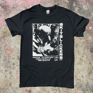 Pode incluir: Camiseta preta com uma estampa gráfica branca de uma banda chamada "Skitslickers". O gráfico inclui uma imagem em preto e branco de uma pessoa, o texto "WARSYSTEM THE LEADER" (sic), "SPRÄCKTA SNUTSKÄLLAR", "ARMED REVOLUTION", "GBG" e "1982".