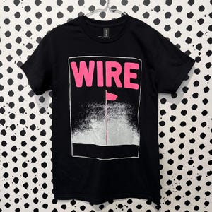May include: A black short-sleeved t-shirt featuring a graphic print with the word WIRE in bold pink letters at the top. Below the text is a white rectangular frame containing a pink flag on a thin pole set against a textured white spray effect.