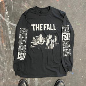 May include: Black long-sleeve shirt featuring the band name "THE FALL" in white block letters. The shirt has a black and white image of the band on the front. The sleeves have a white graphic design.