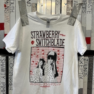 Può includere: T-shirt bianco con la scritta "STRAWBERRY SWITCHBLADE" in bordeaux. Sotto, una grafica di due donne in abiti a pois, incorniciata da una griglia rossa con motivi a fragola. Testo giapponese sottostante.