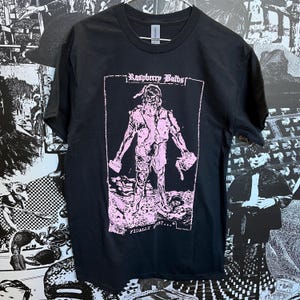 Op de afbeelding: Zwart T-shirt met een roze grafische print van een persoon die twee hamers vasthoudt. De tekst "Raspberry Bulbs" en "FINALLY FIRST..." staat op het shirt gedrukt.