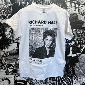 Può includere: Maglietta bianca con un'immagine in bianco e nero di Richard Hell con i Voidoids. Il testo "RICHARD HELL with the Voidoids" e "CALL HELL: (212) 864-0834" è stampato sulla maglietta.