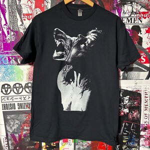 May include: Black t-shirt featuring a white graphic print of a wolf's head with its mouth open and teeth showing. The wolf's head is facing the left side of the image.