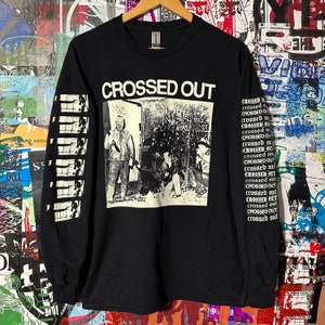 Op de afbeelding: Zwart longsleeve shirt met een witte grafische print van een band genaamd "CROSSED OUT". De grafiek toont een zwart-witfoto van een band voor een gebouw. Het shirt heeft de naam van de band gedrukt op de voorkant en de woorden "CROSSED OUT" herhaald op de linkermouw.