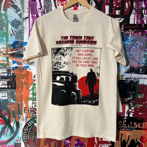 May include: A white t-shirt with a black and white graphic print of a movie poster. The poster says "The Town That Dreaded Sundown: A True Story" and features a car and a person holding an ax. The text on the poster says "Not everyone who comes to this lovers' lane has the same thing on their mind."