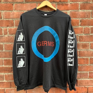 Op de afbeelding: Zwart langarm T-shirt met een blauwe cirkel en het woord "GERMS" in rood. De mouwen hebben zwart-wit afbeeldingen van een persoon.