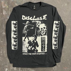 May include: Black long-sleeve shirt with a graphic print. The print features the word "Disclose" above a skull and soldier illustration. The sleeves have a repeating image of a soldier's profile. The text "ONCE THE WAR STARTED....." is at the bottom.