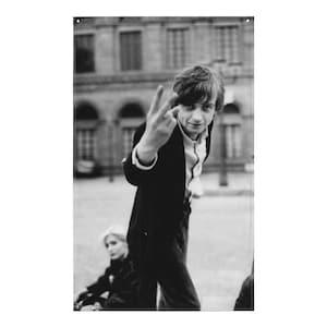 May include: A black and white photo of a man in a suit jacket and white shirt, giving the peace sign with his right hand. He is standing in front of a building with windows.