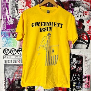 Puede incluir: Camiseta amarilla con un gráfico negro de una persona saltando sobre una bandera con el texto "GOVERNMENT ISSUE" impreso en la camiseta.