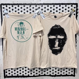 Peut inclure: Un t-shirt à manches courtes de couleur crème avec un motif noir représentant un visage sur le devant et un motif circulaire vert au dos. Le motif au dos représente une silhouette avec le texte MINIMAL-MAN. Le devant inclut le texte MINIMAL MAN.