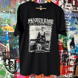Op de afbeelding: Zwart T-shirt met een witte grafische print van een persoon die op een auto zit. De tekst "SHAWTY PIM" en "STILL COMIN' REAL" staat op het shirt gedrukt.
