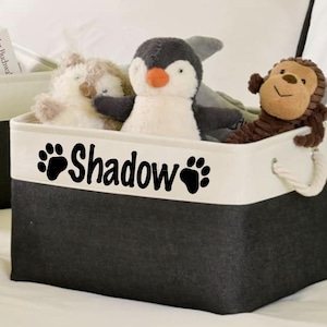 May include: A black and white storage bin with the name "Shadow" and paw prints. The bin is filled with stuffed animals, including a penguin, a monkey, and a fuzzy white animal. The bin has rope handles.