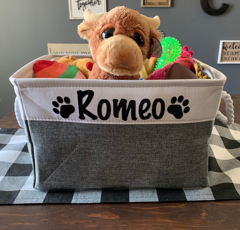 May include: A grey and white canvas storage bin with a rope handle. The bin has a white label with black paw prints and the name "Romeo" on it. The bin is filled with various dog toys, including a plush brown cow, a green squeaky toy, and a red and white striped toy.