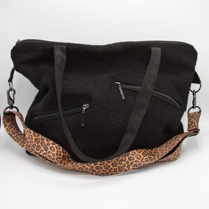 May include: A black textured tote bag featuring two black fabric handles and a detachable shoulder strap with a brown and black leopard print pattern. The front of the bag includes two diagonal zippered pockets with silver-toned metal hardware.