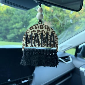 May include: Car mirror hanging ornament with the word "MAMA" in black letters. The ornament has a leopard print design and a black macrame fringe. Two wooden beads are above the ornament, hanging from a black string.