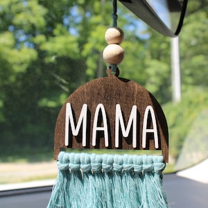 May include: A car rearview mirror hanging ornament featuring a dark brown wooden arch shape with the white text MAMA centered on it. Two natural wooden beads are strung above the arch, and a light blue macrame fringe hangs from the bottom edge.