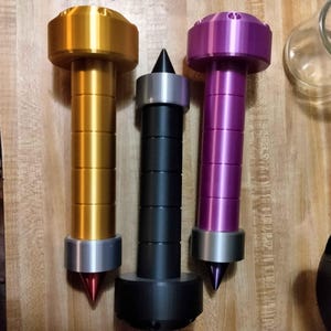 Puede incluir: Tres pies de componentes de audio metálicos en dorado, negro y morado. Cada pie tiene un cuerpo cilíndrico con una punta cónica y una parte superior circular. El pie dorado tiene una punta roja, el pie negro tiene una punta negra y el pie morado tiene una punta morada.